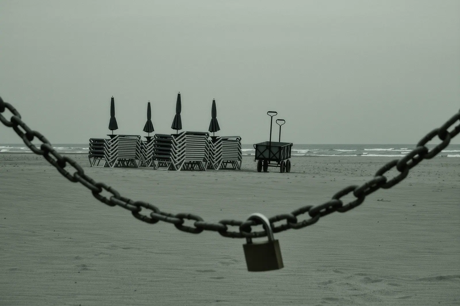 Stacked sun loungers, folded parasols and a beach cart chained together on the sand, the heavy chain and padlock close in the foreground.