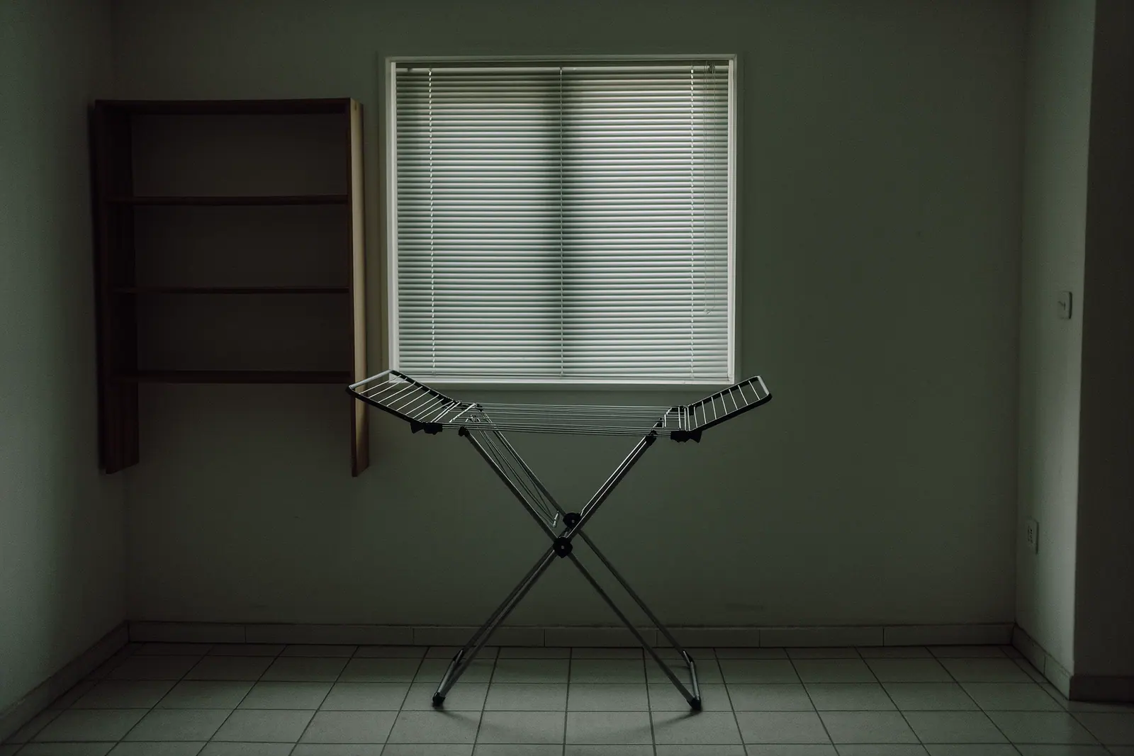 A plain long-let flat interior with a folding drying rack standing open on the tiled floor in the foreground, a shuttered window and empty shelves behind.