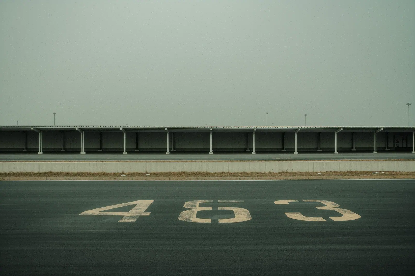 An empty transfer pickup bay outside an airport terminal, large worn painted bay numbers filling the tarmac in the foreground, a bare kerb and plain shelter behind.