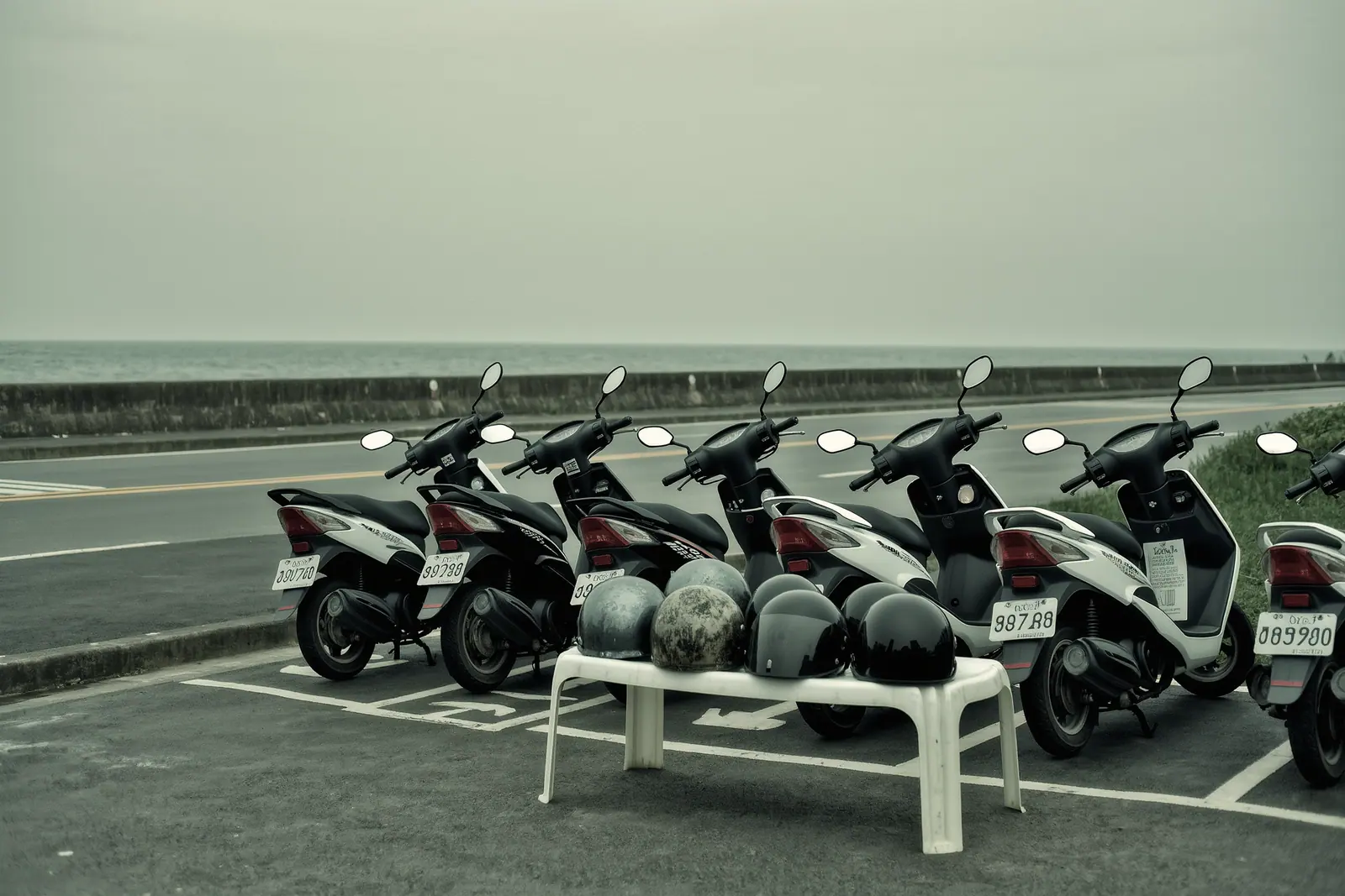 Scooters parked nose-in inside a marked rental bay beside a coastal road, a stack of scuffed helmets on a white plastic table in the foreground.