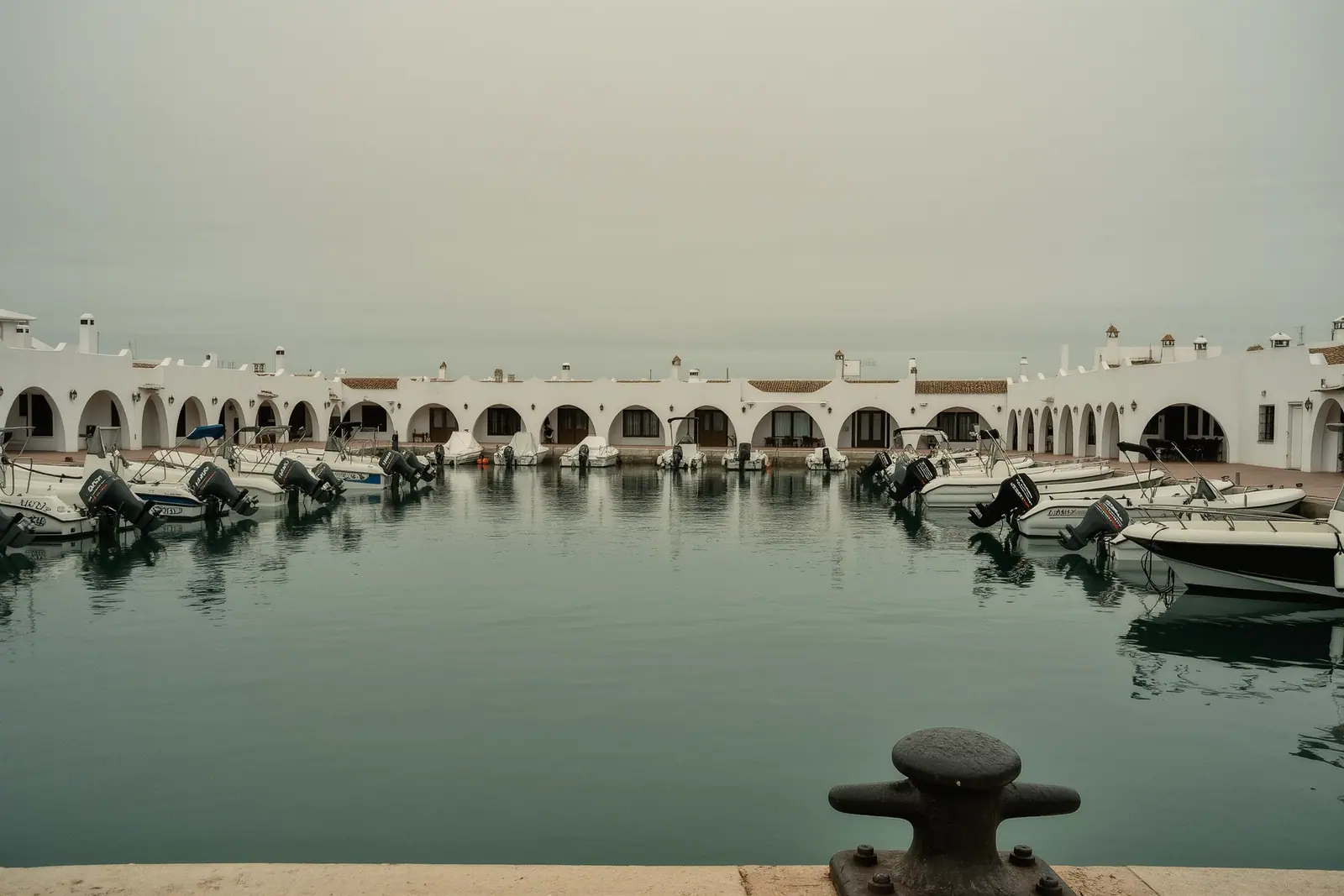 The inner basin at Puerto Marina with small motorboats tied along finger pontoons, a mooring cleat and the pontoon edge in the foreground, whitewashed arched buildings around the water.