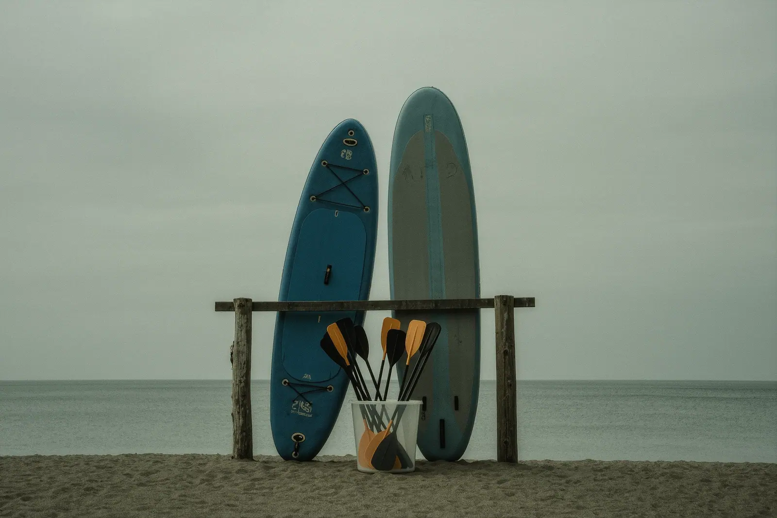 Paddleboards standing on edge in a weathered wooden rack on the beach, paddles standing in a plastic barrel in the foreground, flat grey sea behind.