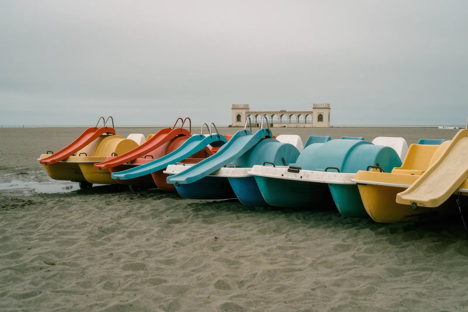 Pastel plastic pedal boats with slides hauled up on the sand at Benalmádena, the whitewashed arches of Puerto Marina behind.