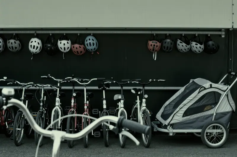 Children's bicycles and a child trailer standing in a metal rental rack, helmets of several sizes hanging on a rail behind them.